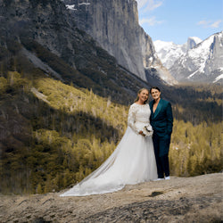 Bride in a long-sleeved lace wedding gown and groom in a dark suit from Addicted Bespoken posing together on a mountain overlook with snowy peaks in the background