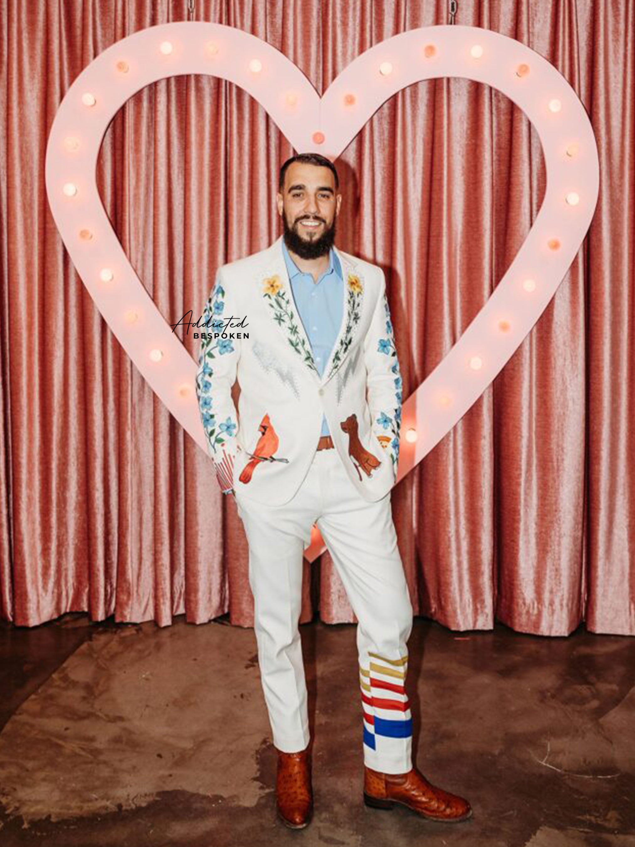 Man wearing a bespoke white embroidered suit with colorful bird and stripe motifs from Addicted Bespoken, standing in front of a heart-shaped light backdrop