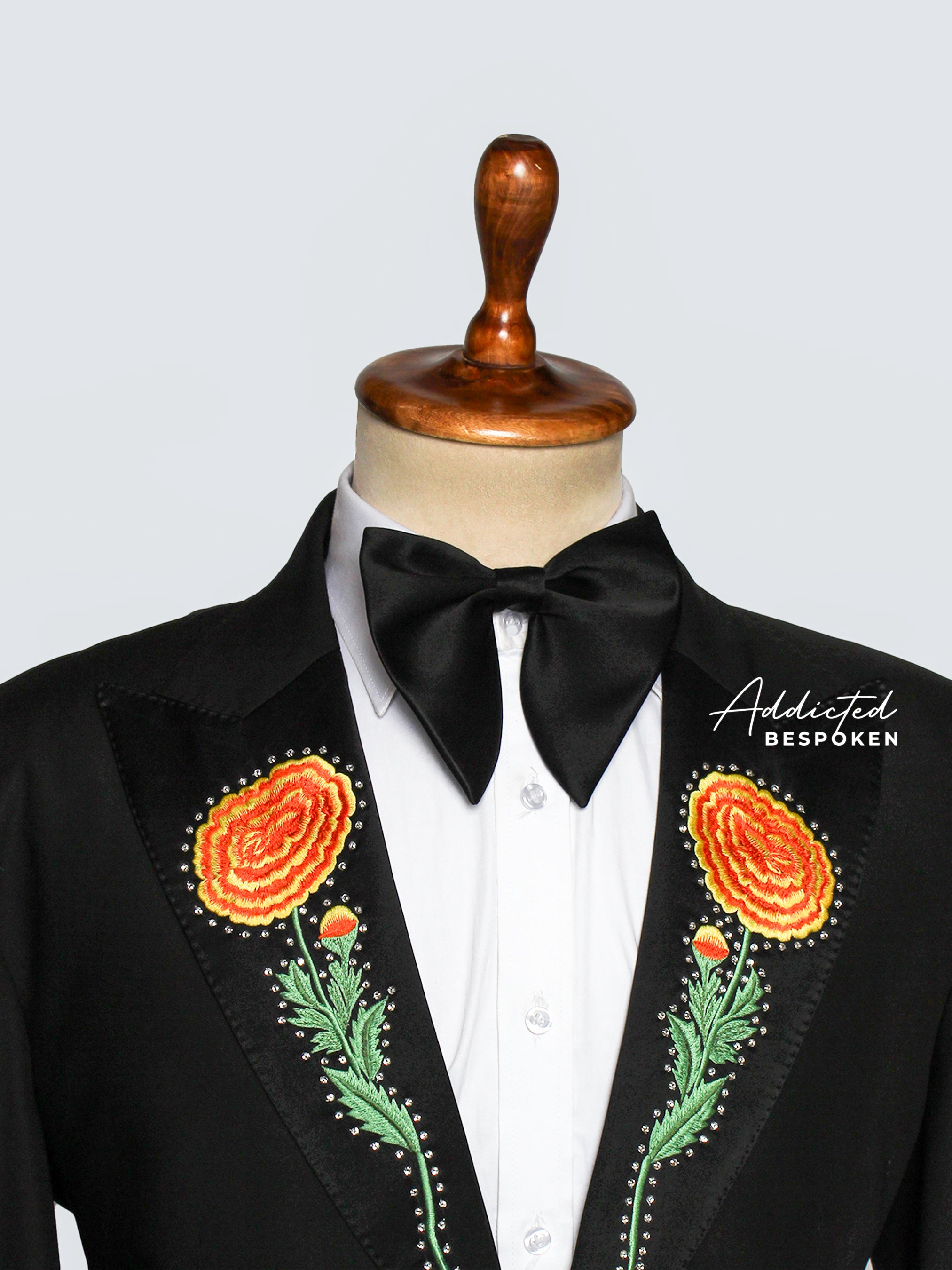Close-up of a Black tuxedo with floral embroidery and a black bow tie against a light gray background on a mannequin.