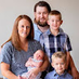 Smiling family portrait of two parents with three young children including a baby, posing together indoors against a neutral background