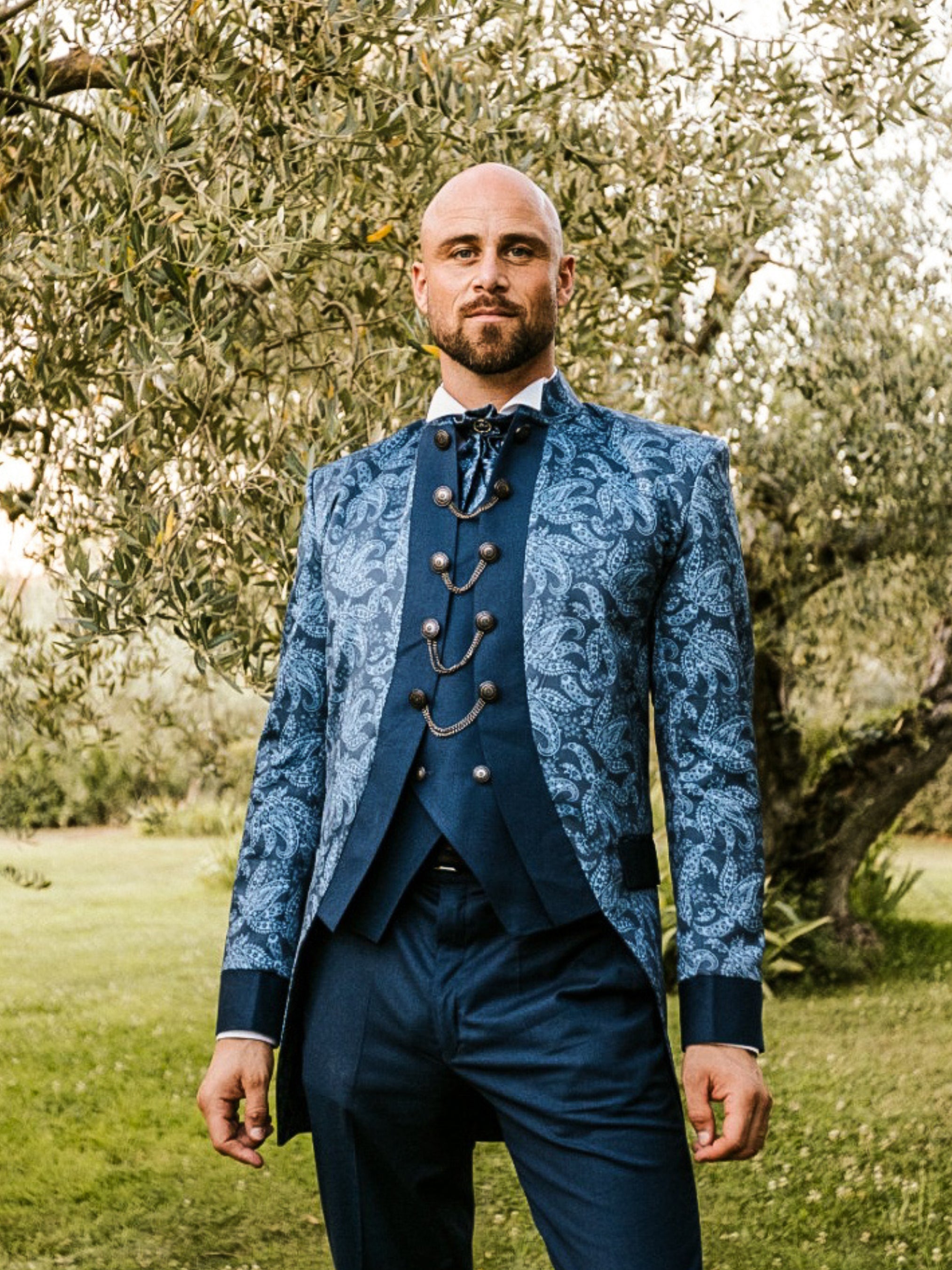 Man wearing an ornate blue brocade three-piece suit with chain closures from Addicted Bespoken, posing outdoors in a garden setting.