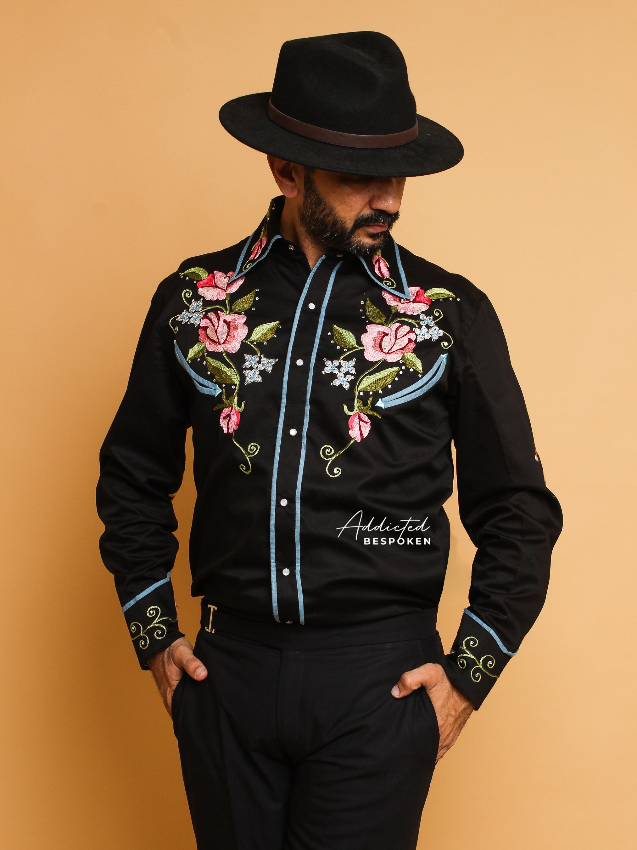 Man wearing a black western-style embroidered shirt with colorful floral motifs from Addicted Bespoken, styled with black hat and trousers.