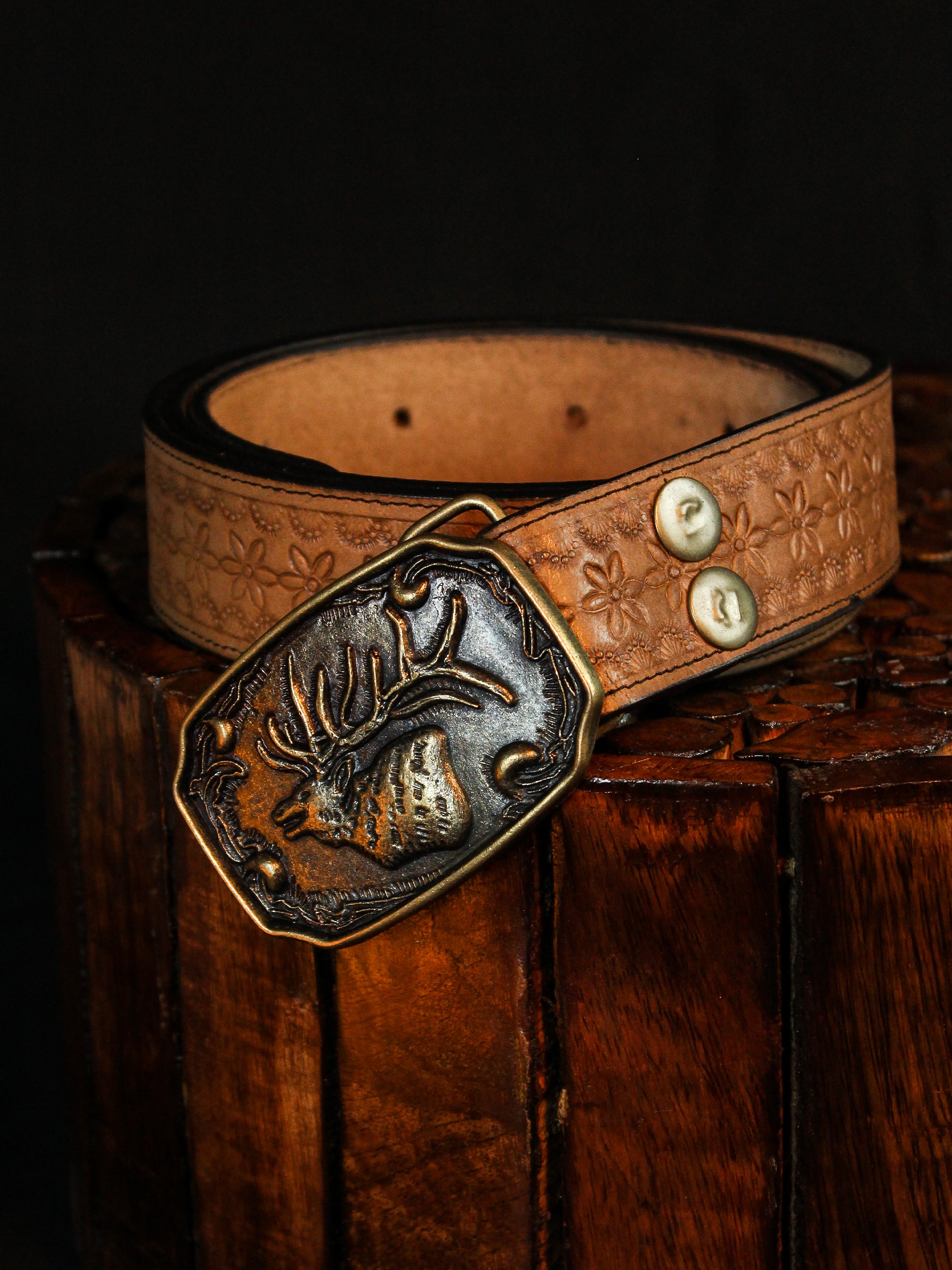 Handcrafted tan leather belt with embossed pattern and antique brass deer head buckle from Addicted Bespoken displayed on a wooden surface.