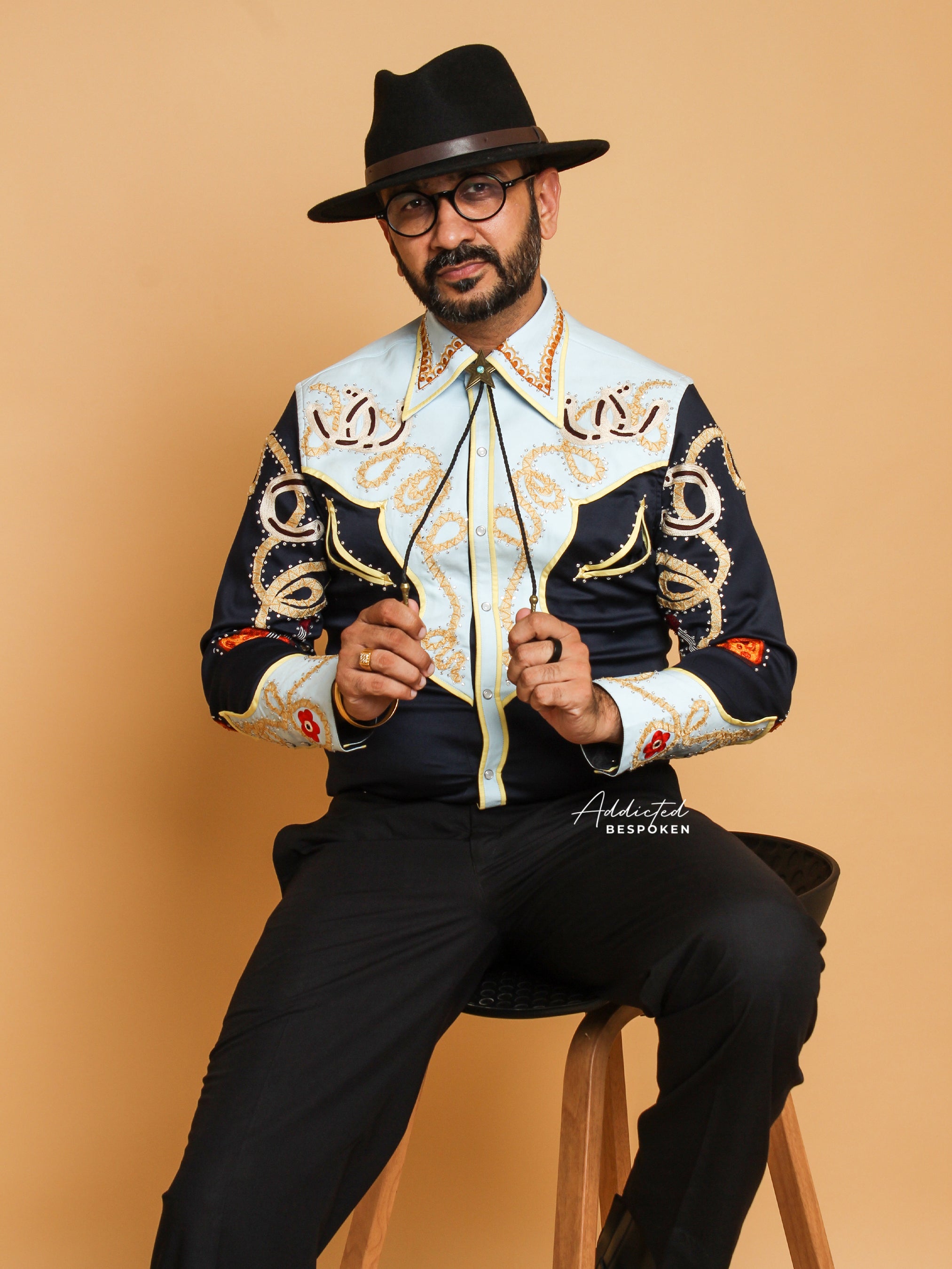 A person wearing a statement shirt with gold and orange embroidery on a navy background, paired with black trousers and black hat.