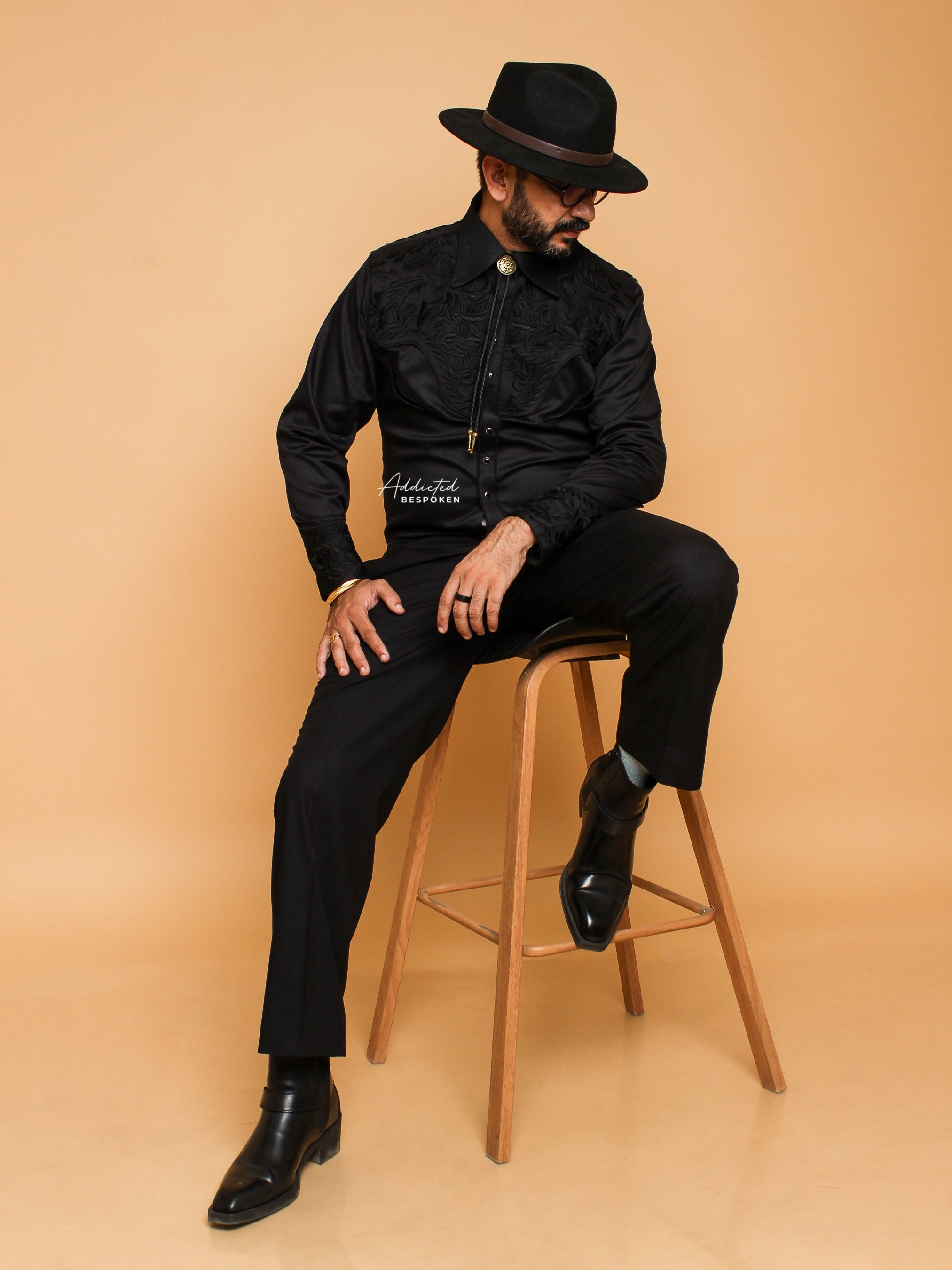 A man sitting on a stool, wearing a black embroidered shirt with long sleeves, tailored black trousers, and polished leather boots. He has on a classic black fedora hat and a distinctive bolo tie featuring a gold medallion.