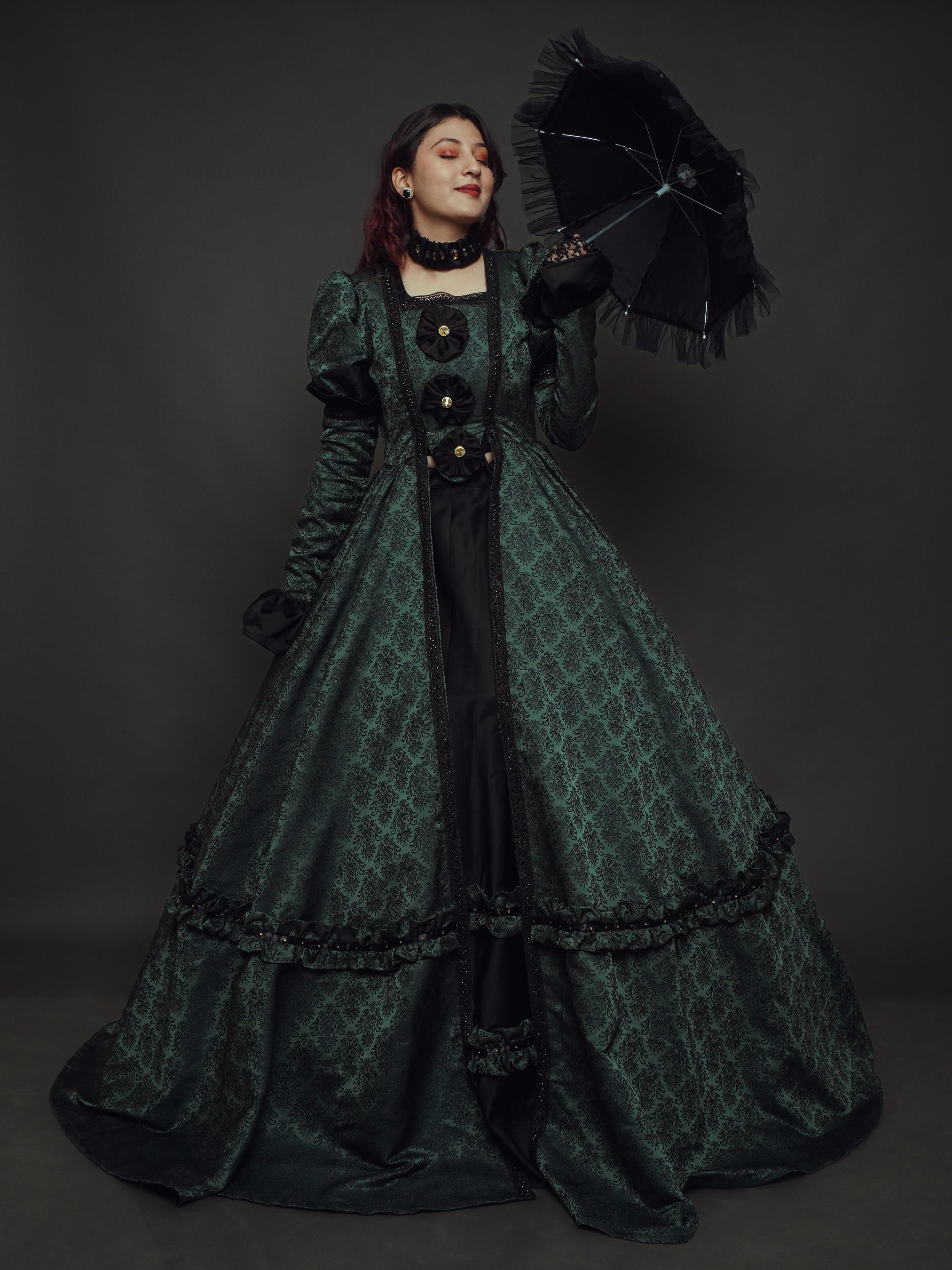 A woman posing in a luxurious green brocade gown with black satin accents, featuring a flowing silhouette and delicate details.
