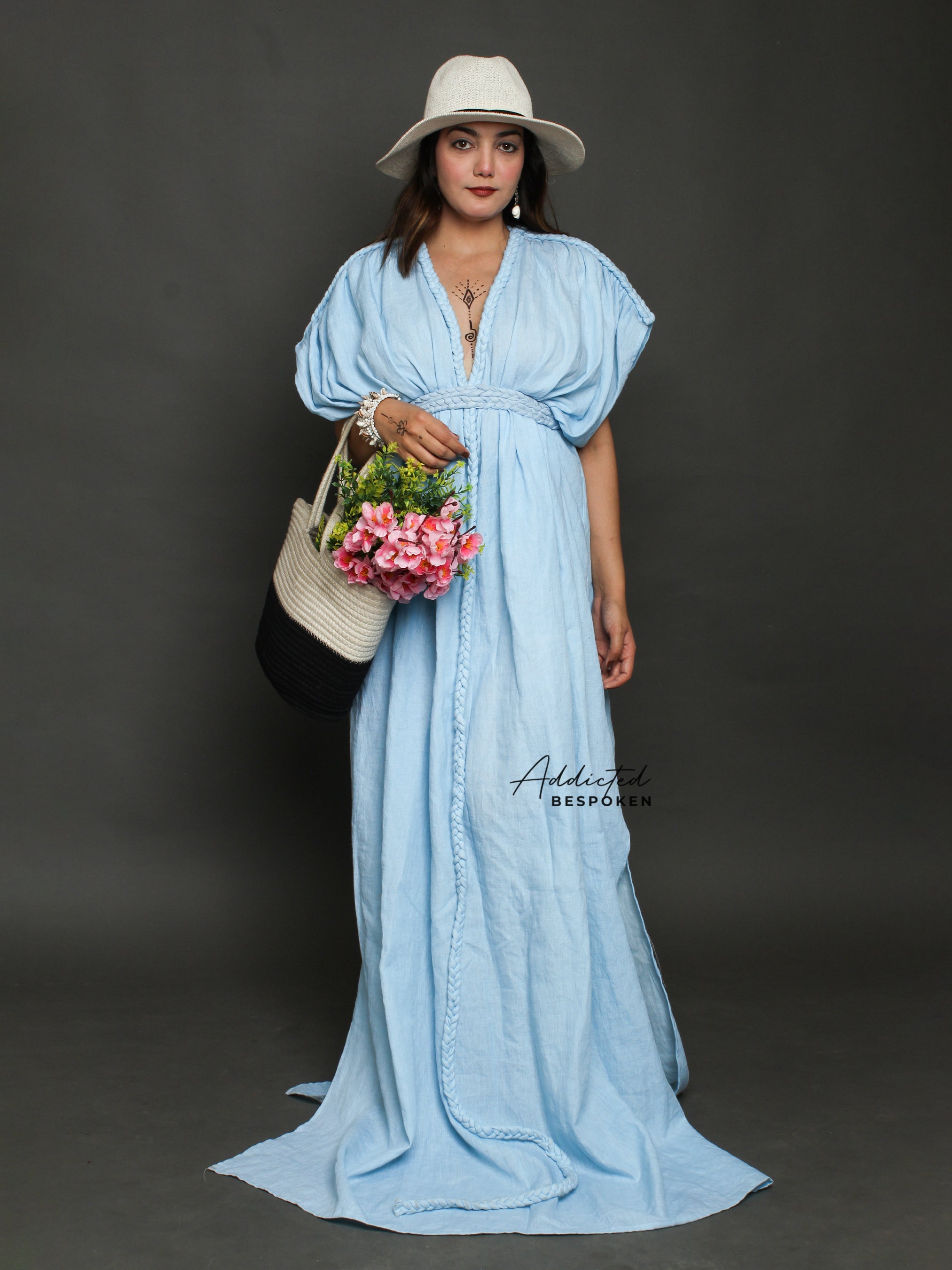 A woman wearing a blue linen boho style baggy dress with a deep V neck, short sleeves, and long length. She is also wearing a wide brim hat and carrying a bag with flowers.