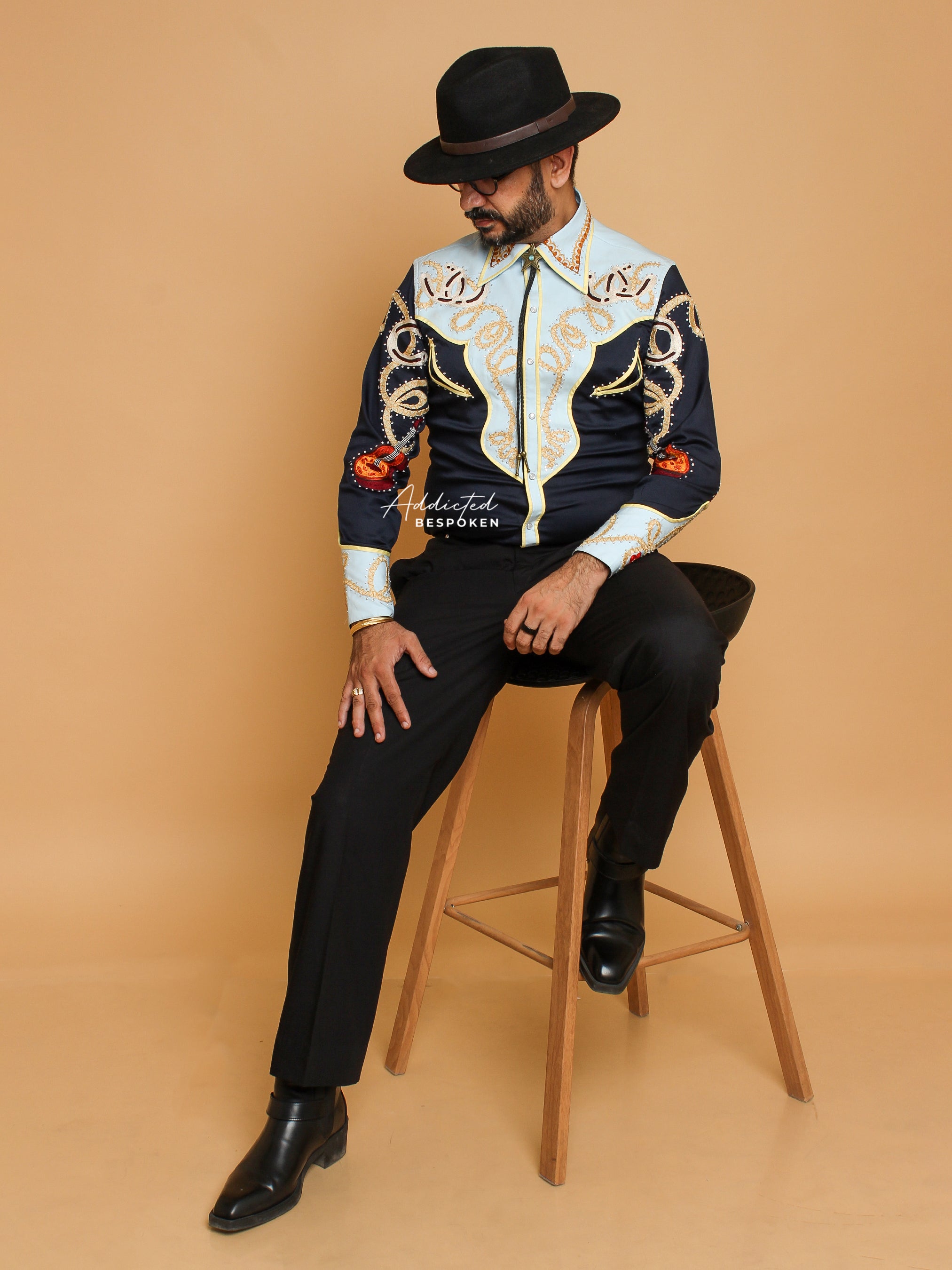 A person wearing a deep navy shirt with gold and orange intricate embroidery, tailored black trousers, and black boots, sitting on a wooden stool against a beige background.