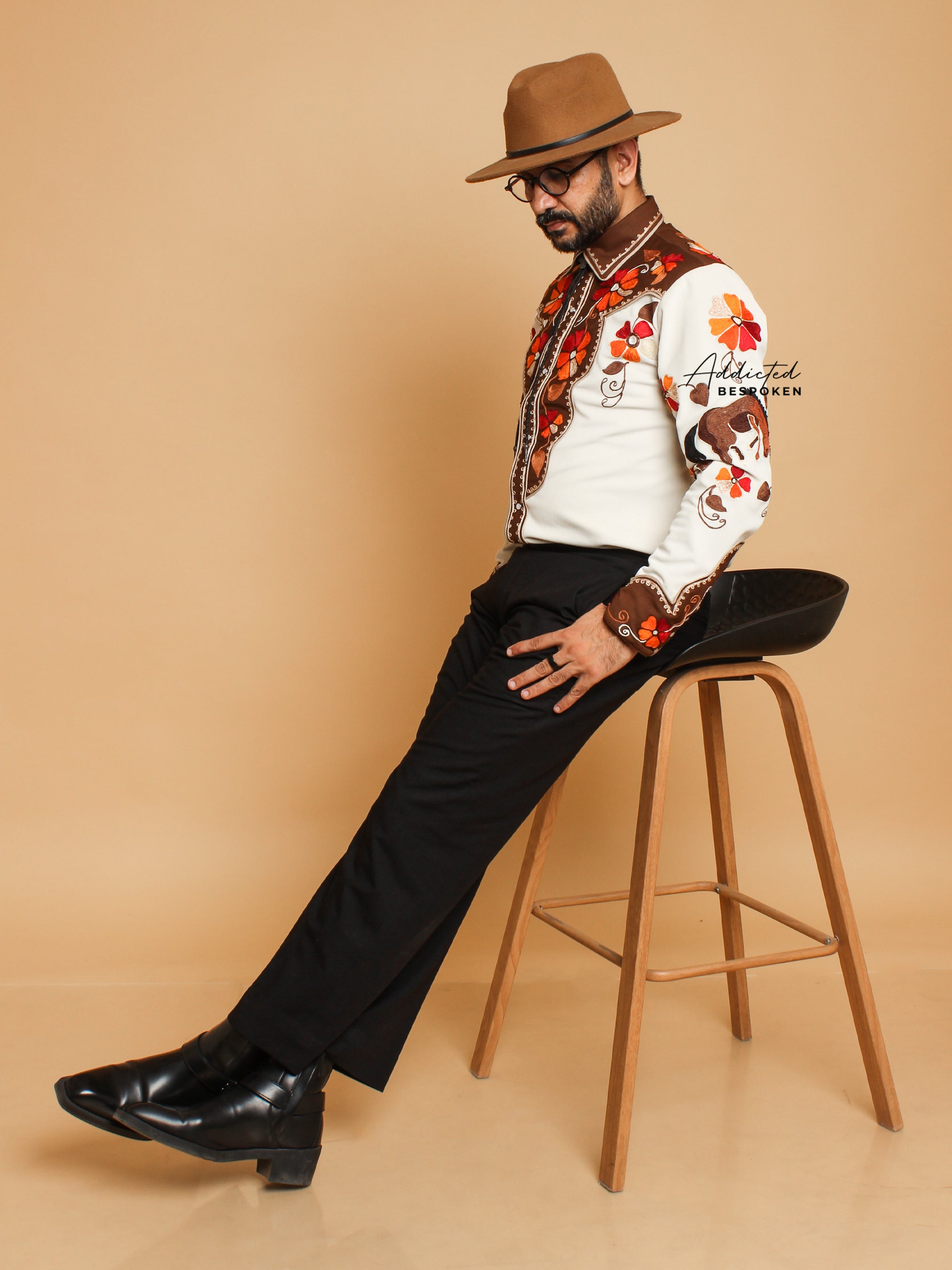 Seated angled view of male model wearing cream embroidered western shirt with hat โ Addicted Bespoken