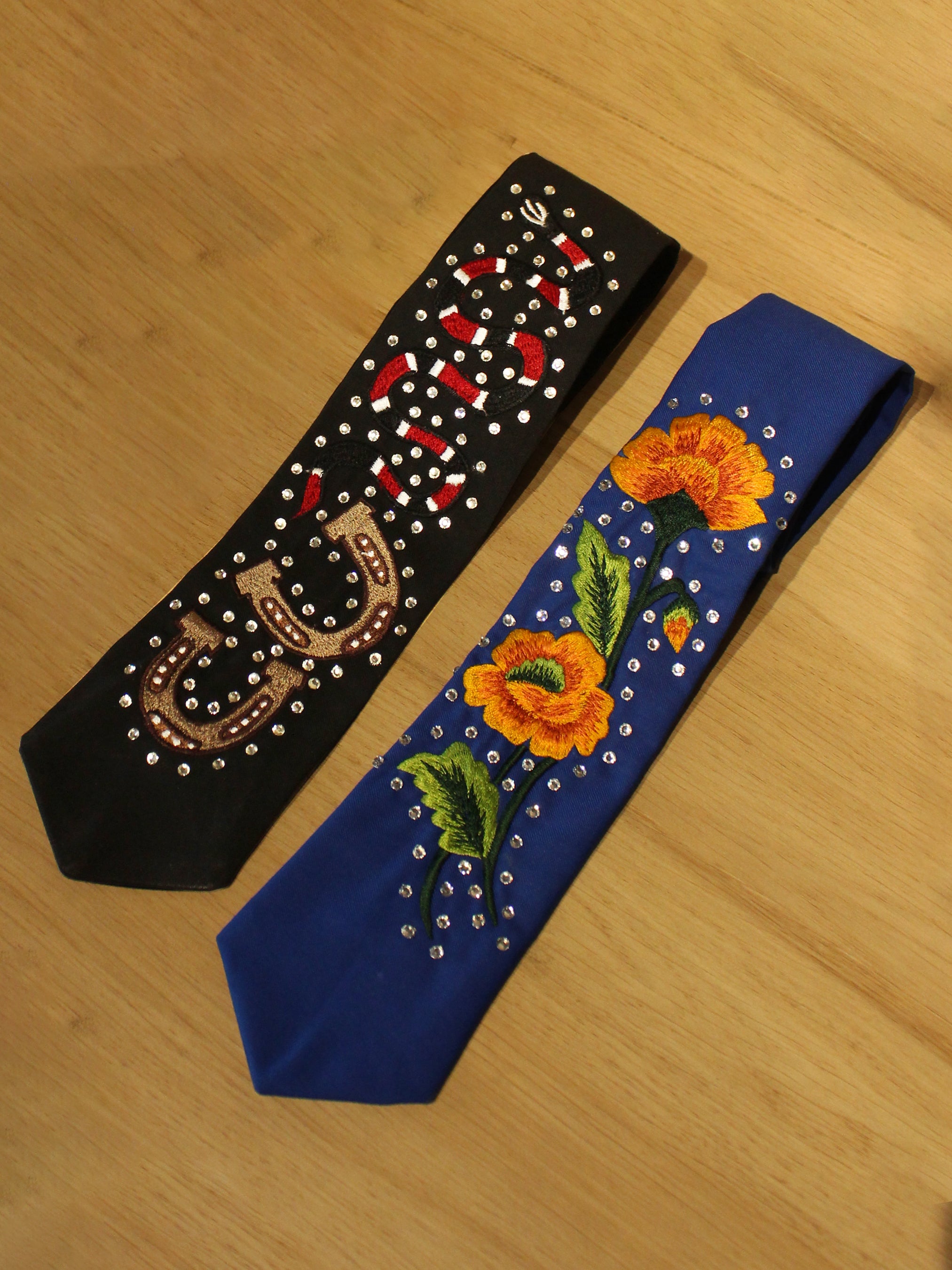 Two embroidered neckties from Addicted Bespoken – one black with rhinestones and horseshoe motif, one blue with vibrant floral embroidery displayed on wooden surface