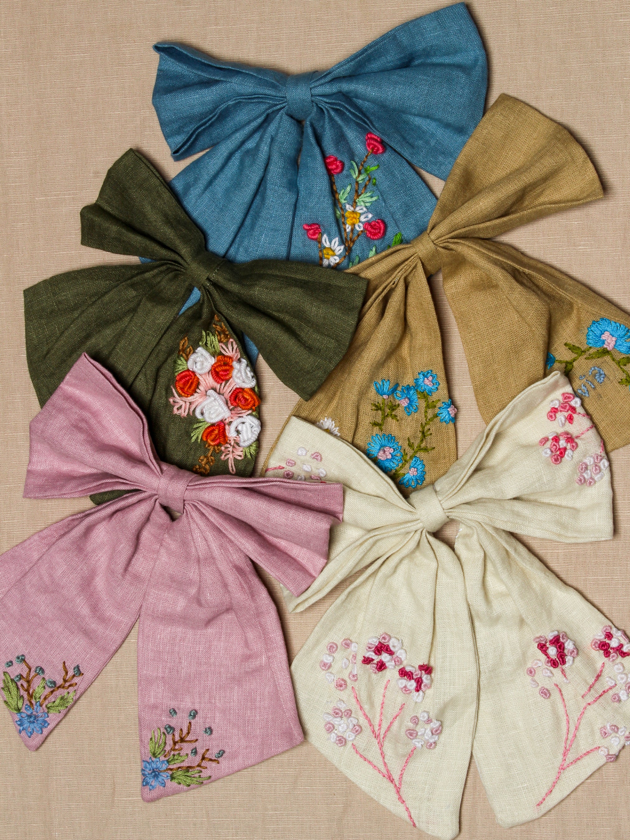 Set of large embroidered fabric bows from Addicted Bespoken in blue, olive, gold, pink and cream with floral detailing displayed on a neutral background