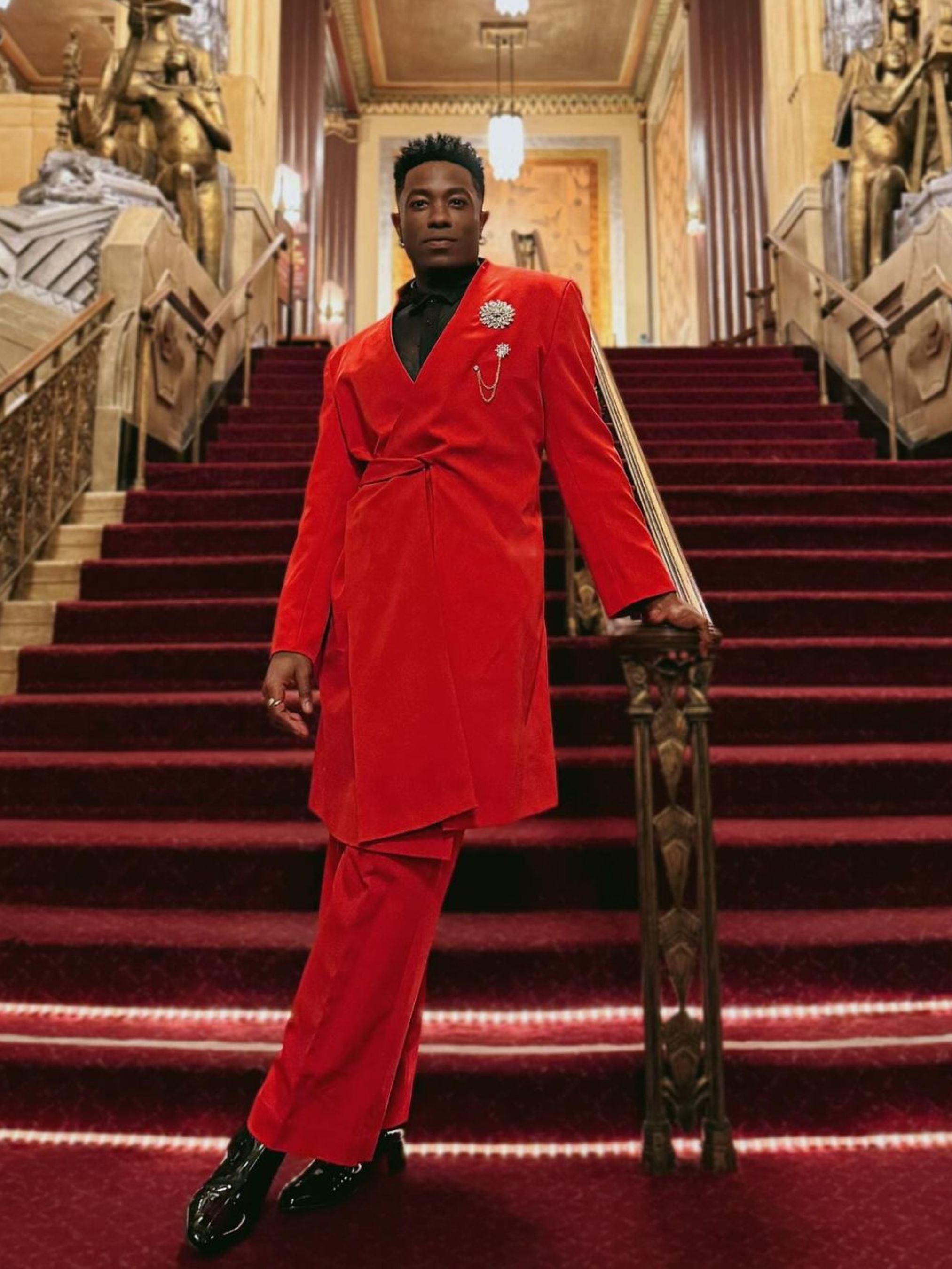 Man wearing a bold red bespoke suit from Addicted Bespoken with brooch detail, standing on an ornate grand staircase in a luxurious interior