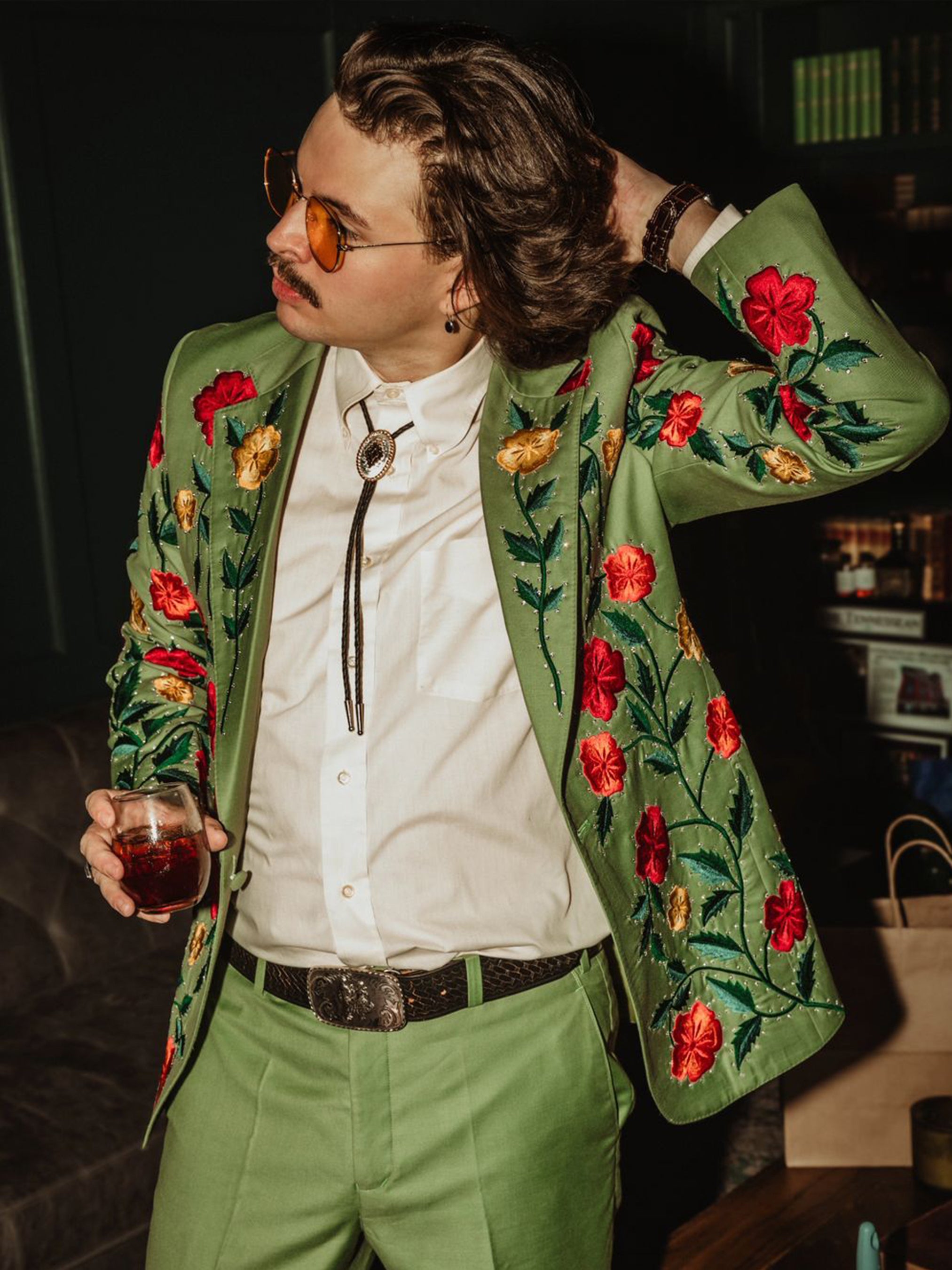Man wearing a green bespoke suit jacket richly embroidered with red and yellow flowers from Addicted Bespoken, styled with bolo tie and sunglasses holding a drink