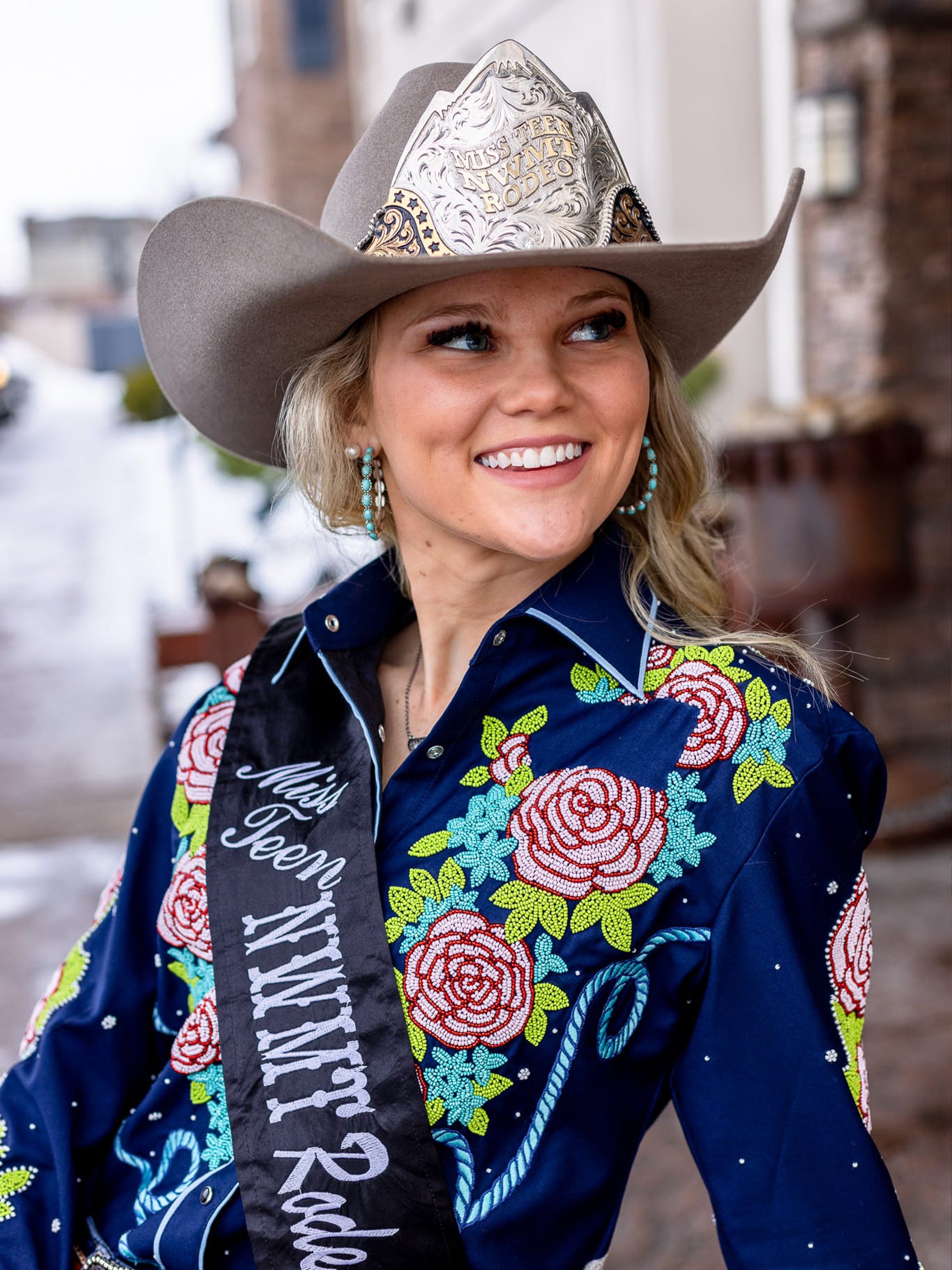 Young woman in a grey cowboy hat wearing a navy western shirt richly embroidered with pink roses from Addicted Bespoken, smiling while wearing a rodeo sash.