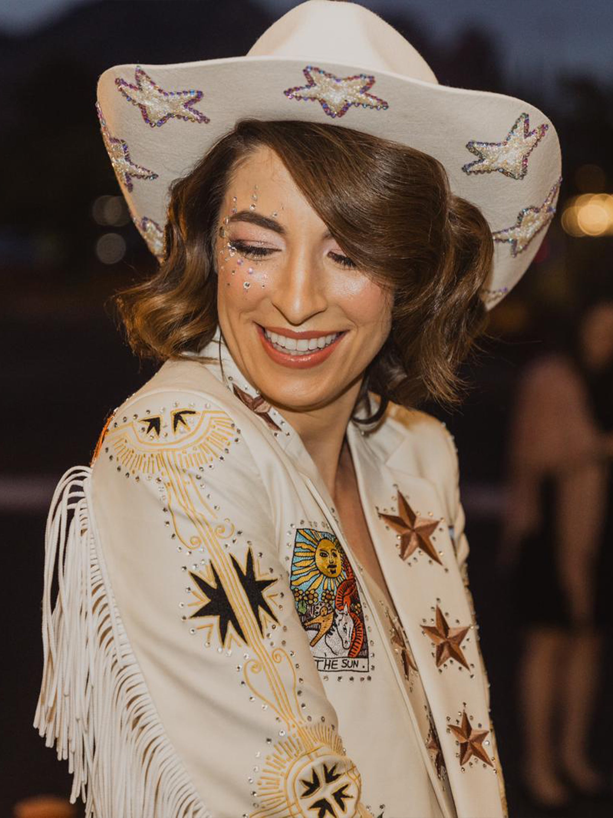Woman wearing a white western-style fringed jacket with celestial star embroidery and matching cowboy hat from Addicted Bespoken, smiling at an event.